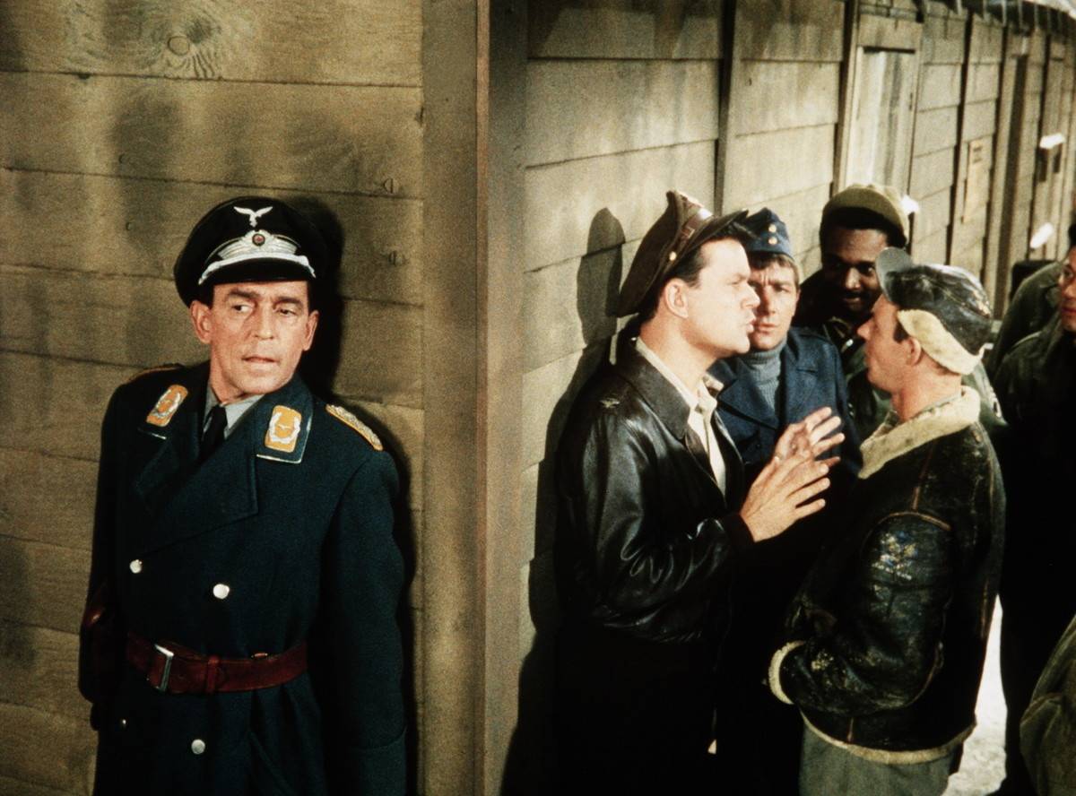 Hogan's Heroes main characters are speaking while a guard is pressed around the wall behind a corner nearby