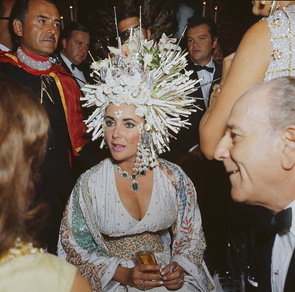 elizabeth taylor wearing an elaborate headdress of pearls and fake flowers, a jewelled dress and an emerald necklace